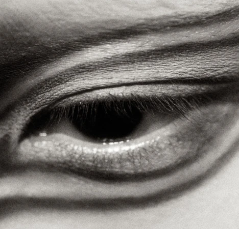 Close-up black and white photograph of a human eye with visible eyelashes, eyebrow and skin texture, showcasing a natural expression.