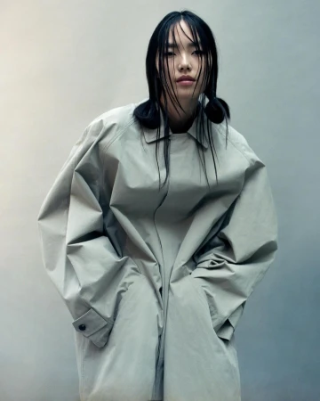 Asian woman with wet dark hair wearing an oversized grey trench coat against a textured neutral background by Myrthe Giesbers
