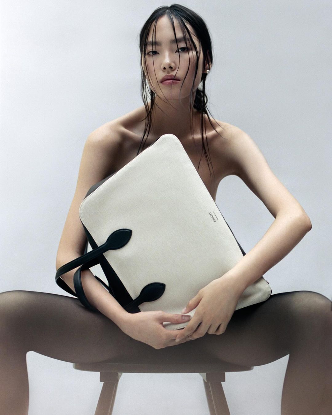 Elegant portrait of a young Asian woman sitting on a stool, posing with a large black and white tote bag in a bright studio setting.