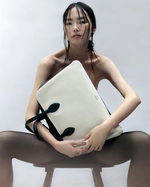 Elegant portrait of a young Asian woman sitting on a stool, posing with a large black and white tote bag in a bright studio setting.