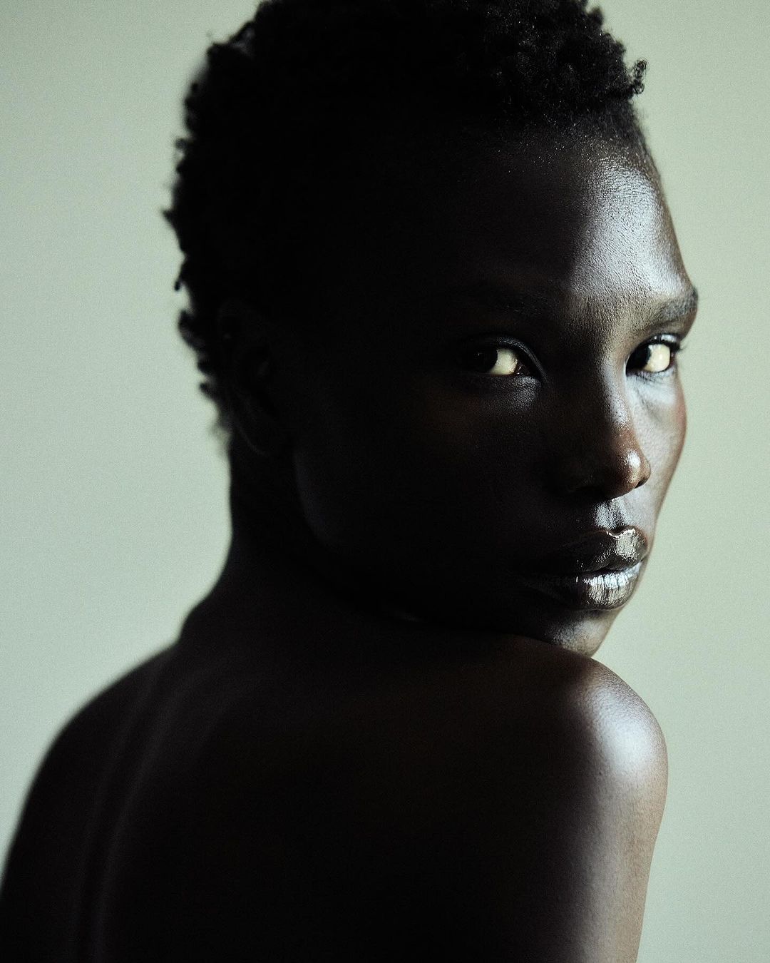 Portrait of an adult Black woman with dark, glossy skin, short hair, and intense gaze, against a neutral backdrop. Minimalist beauty shot.