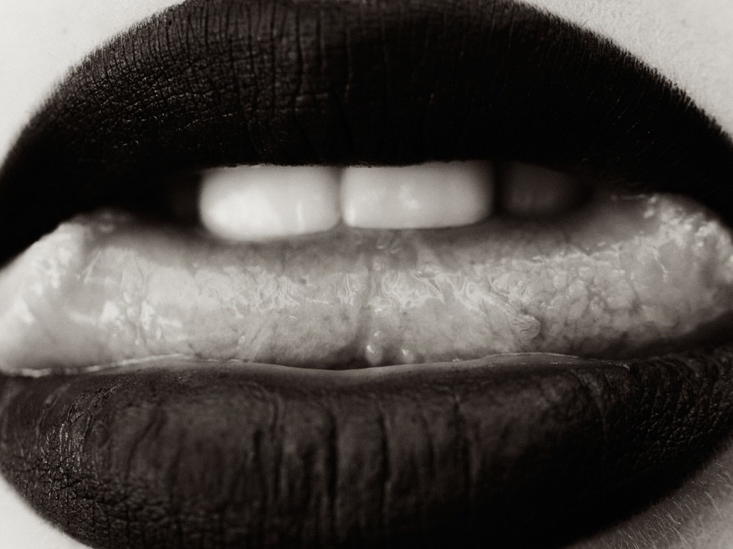 Extreme close-up of open mouth with red lipstick, white teeth, and extended tongue showing texture in a monochrome studio shot.