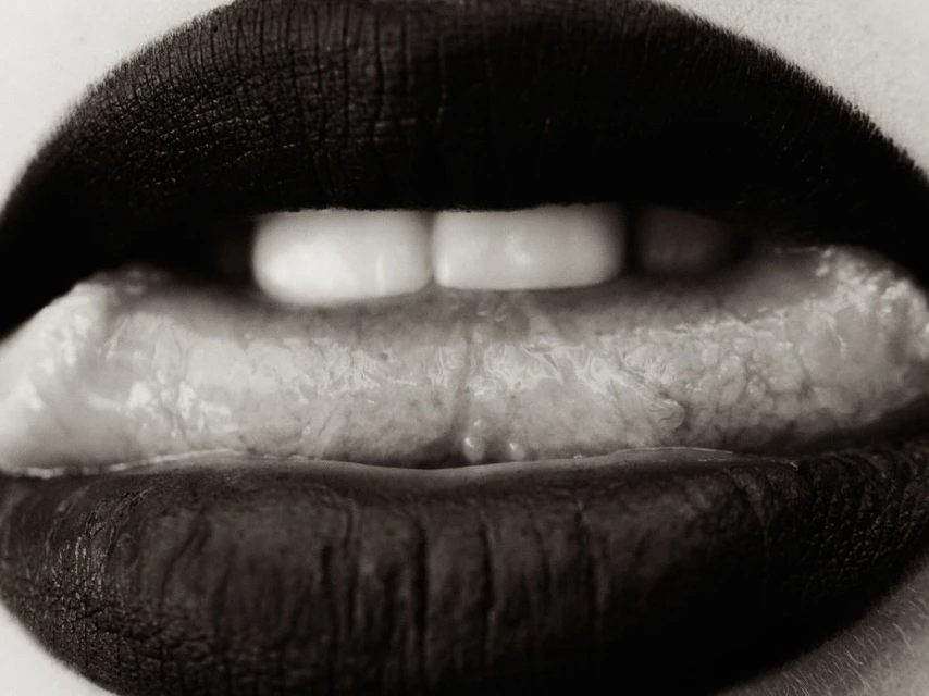Extreme close-up of open mouth with red lipstick, white teeth, and extended tongue showing texture in a monochrome studio shot.