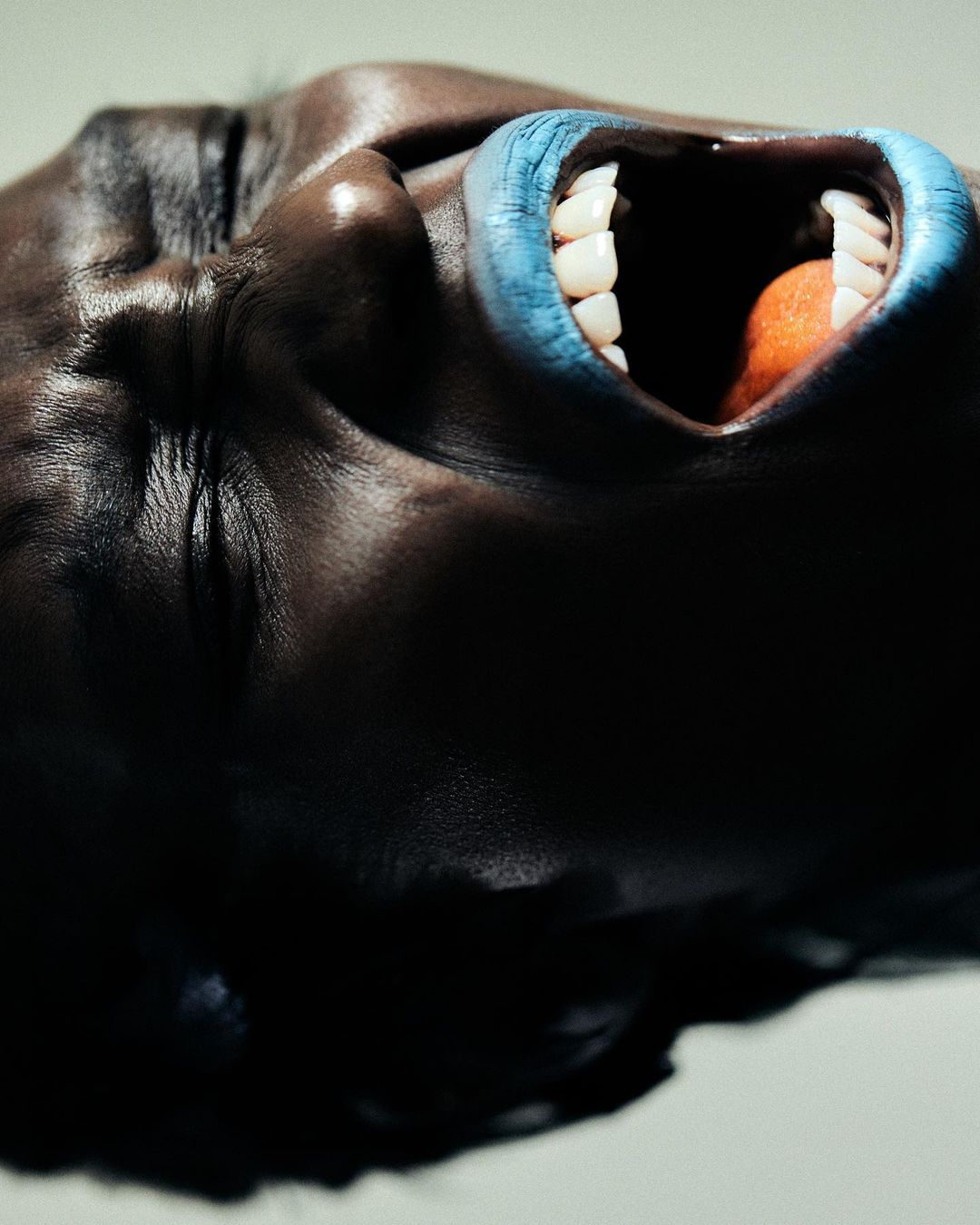 Close-up studio shot of a Black adult female's face with an open mouth, teeth, and an orange object in her mouth; blue lipstick