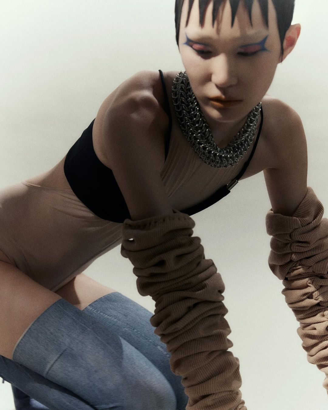 A young adult female model wearing a beige bodysuit, black bra top, light blue jeans, and a chain necklace poses in a studio.