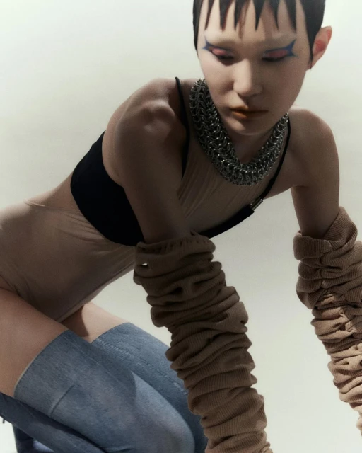 A young adult female model wearing a beige bodysuit, black bra top, light blue jeans, and a chain necklace poses in a studio.