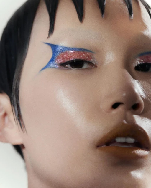 Close-up beauty shot of a person with short dark hair and creative colorful makeup around the eyes and lips against a plain background.