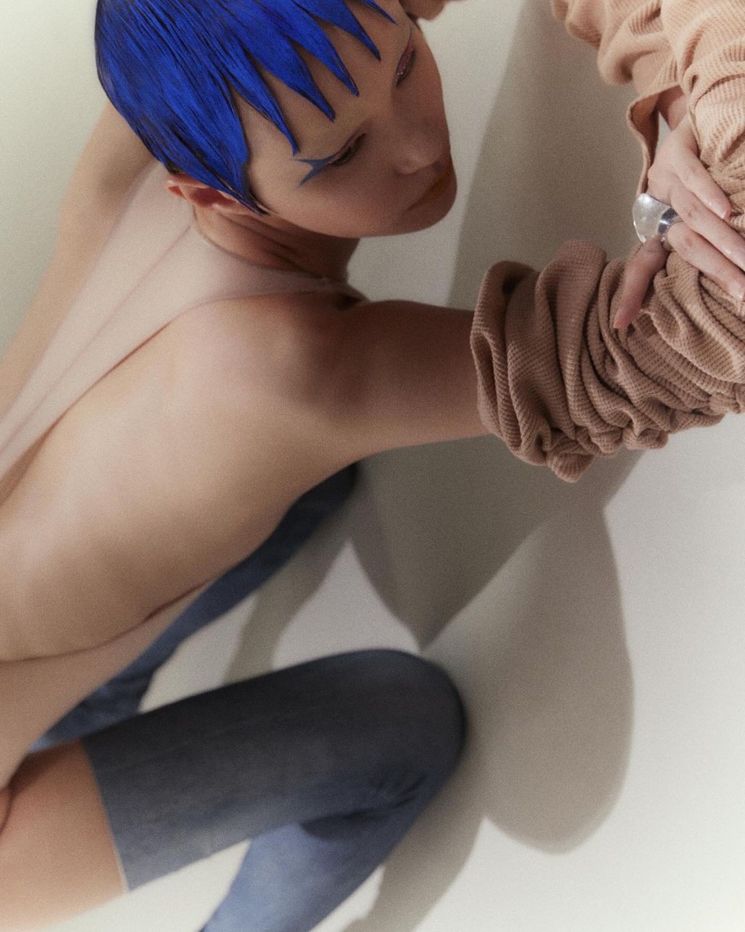 Person with vibrant blue hair and makeup strikes a pose, showcasing neutral top and blue bottoms against a muted white background.
