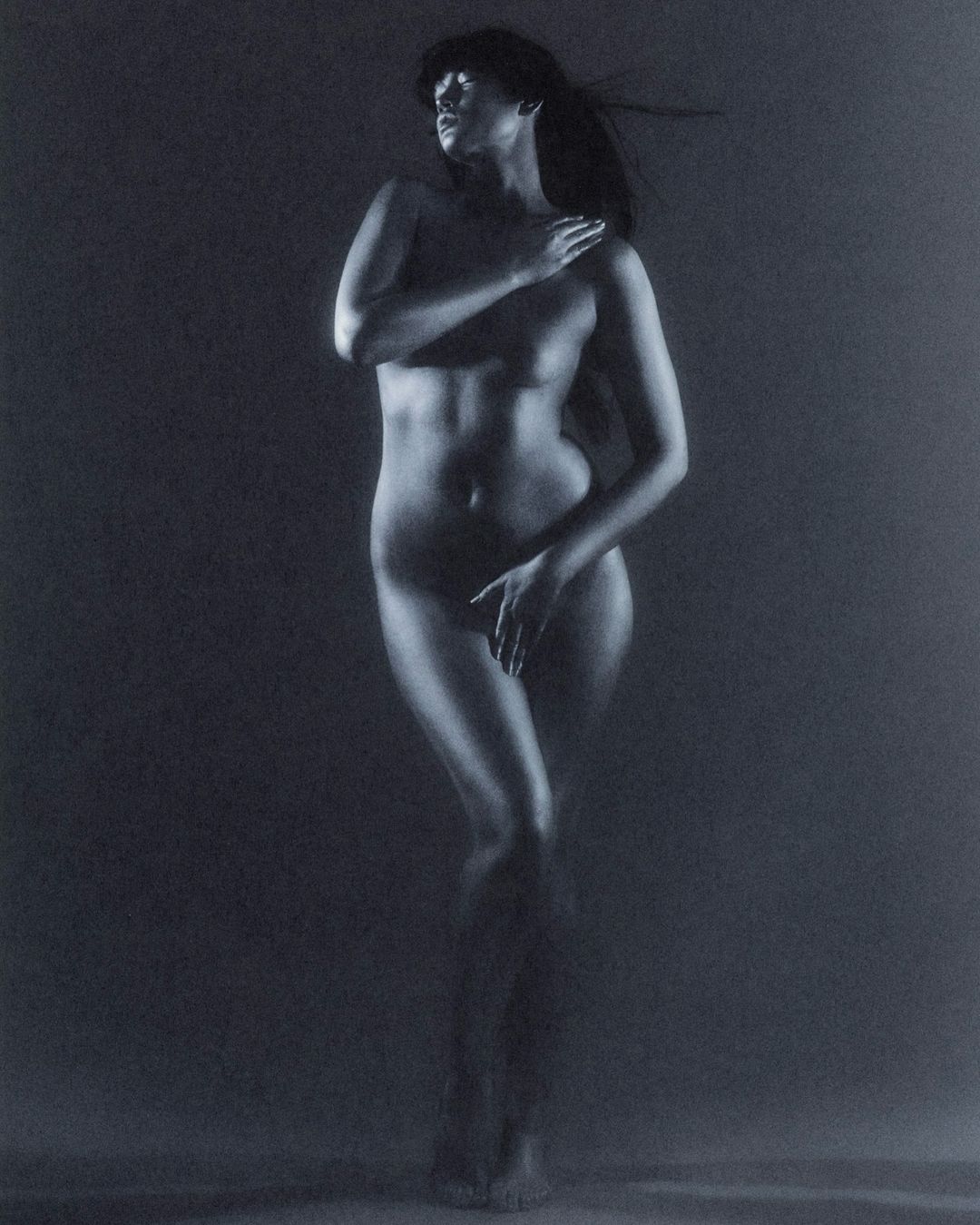 Nude woman stands illuminated in a dark setting, covering her chest and groin with her hands, exuding artistic elegance.