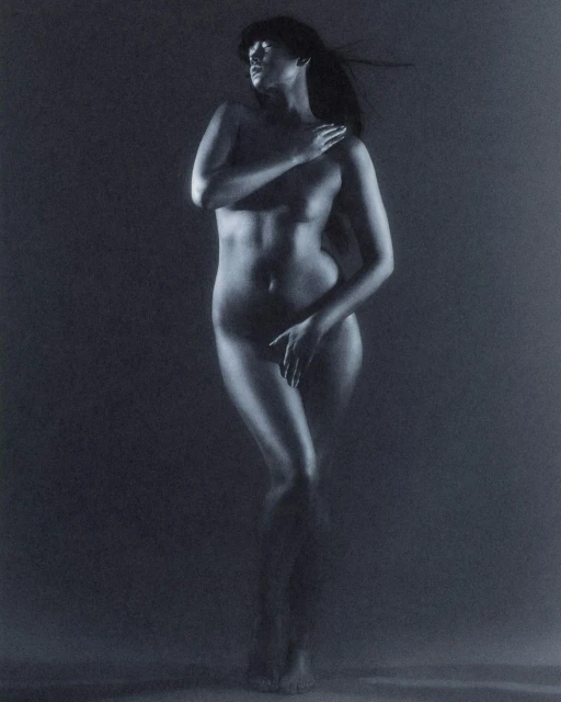 Nude woman stands illuminated in a dark setting, covering her chest and groin with her hands, exuding artistic elegance.