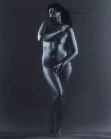 Nude woman stands illuminated in a dark setting, covering her chest and groin with her hands, exuding artistic elegance.