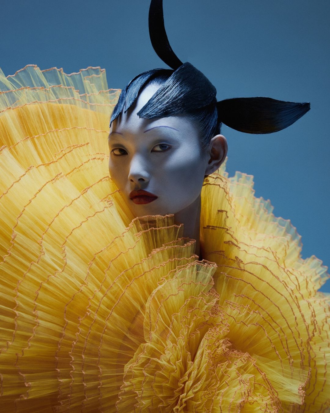 Woman in elaborate yellow ruffled dress and black sculptural headpiece against a blue background in a beauty and fashion studio shot.