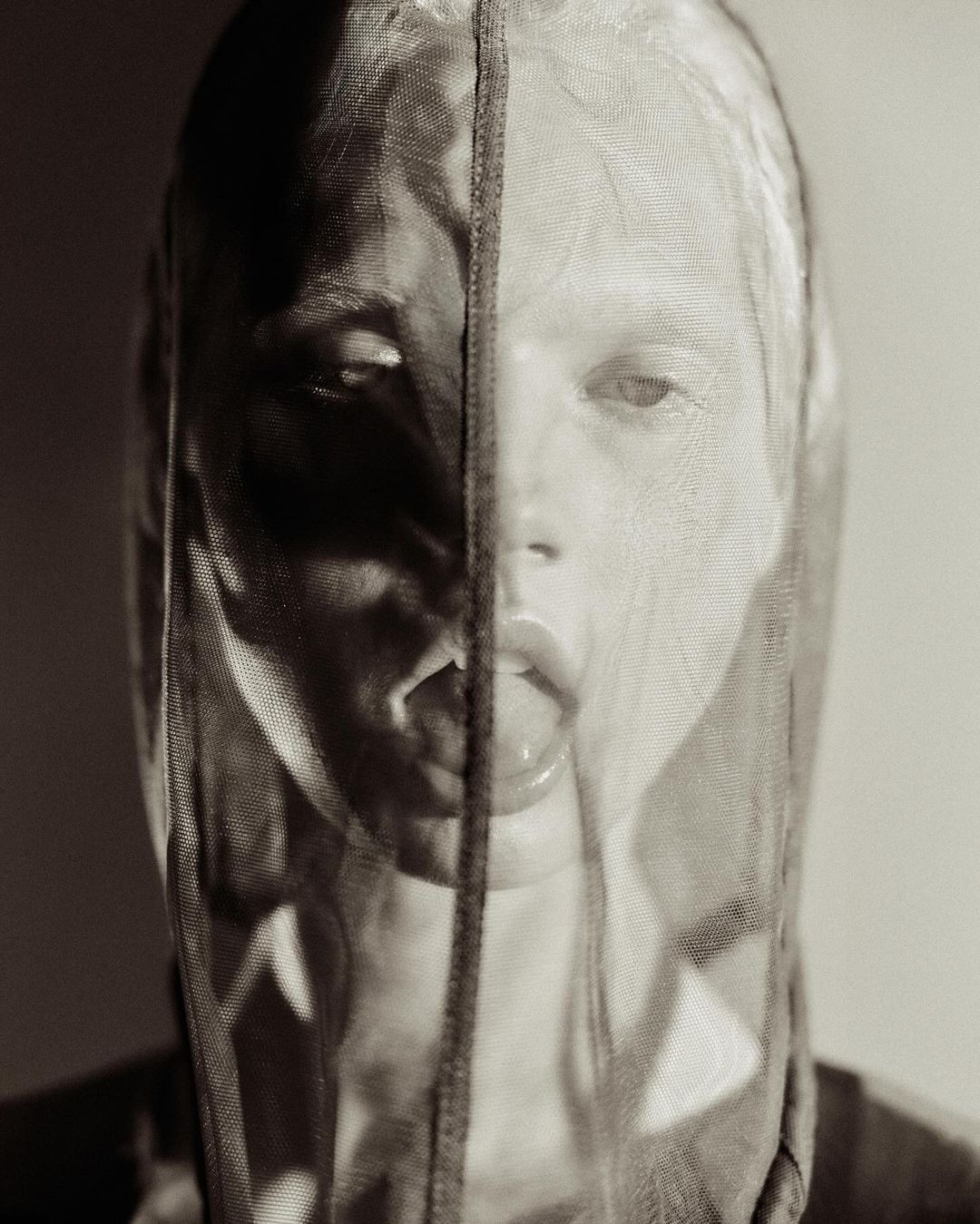 Striking monochrome portrait of an adult woman with her tongue out, partially concealed behind a sheer net fabric in studio lighting.