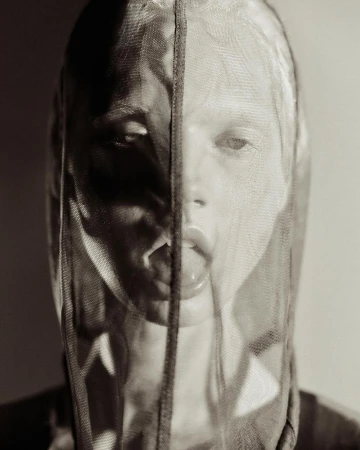 Striking monochrome portrait of an adult woman with her tongue out, partially concealed behind a sheer net fabric in studio lighting.