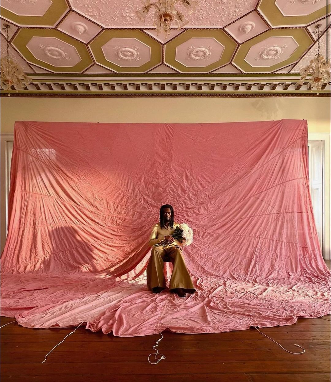 Person in gold outfit seated with flowers against pink backdrop, ornate ceiling and chandelier, captured with an eye-level view.