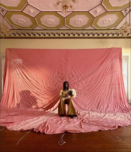 Person in gold outfit seated with flowers against pink backdrop, ornate ceiling and chandelier, captured with an eye-level view.