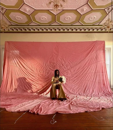 Person in gold outfit seated with flowers against pink backdrop, ornate ceiling and chandelier, captured with an eye-level view.