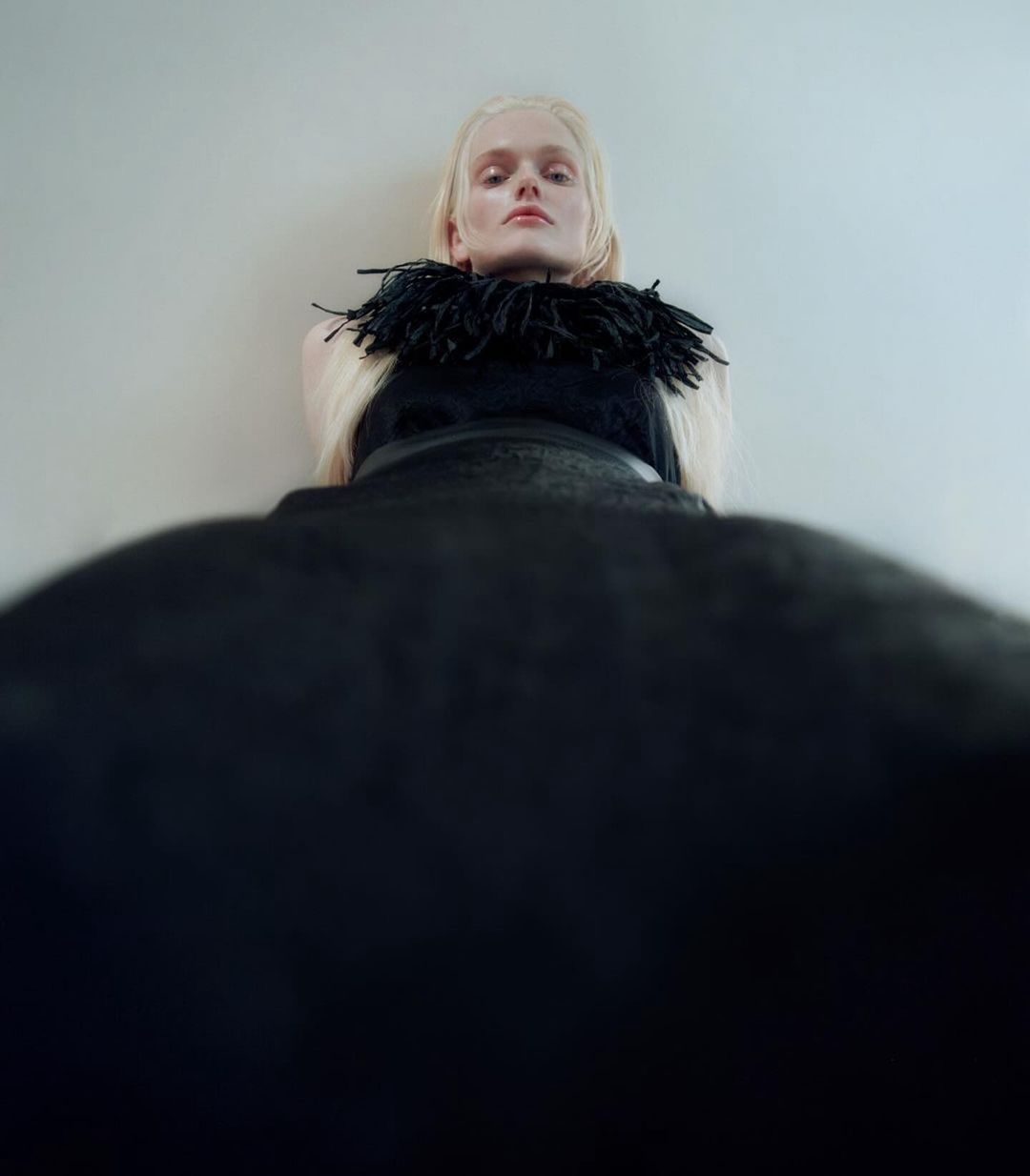 Fashion shot of an adult female model with pale blonde hair wearing a black dress with a feathered collar and standing against a wall.