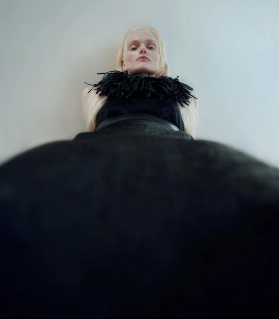 Fashion shot of an adult female model with pale blonde hair wearing a black dress with a feathered collar and standing against a wall.
