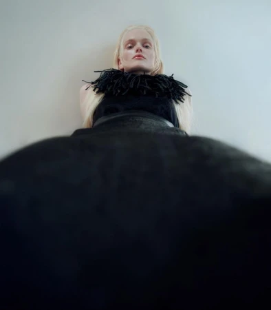 Fashion shot of an adult female model with pale blonde hair wearing a black dress with a feathered collar and standing against a wall.