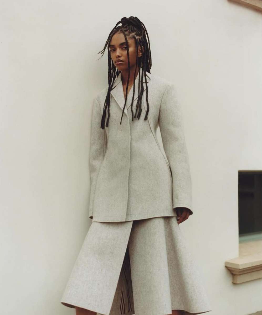 Fashion portrait of a Black woman with braided hair, wearing a neutral blazer and culottes, posing against a textured off-white wall.