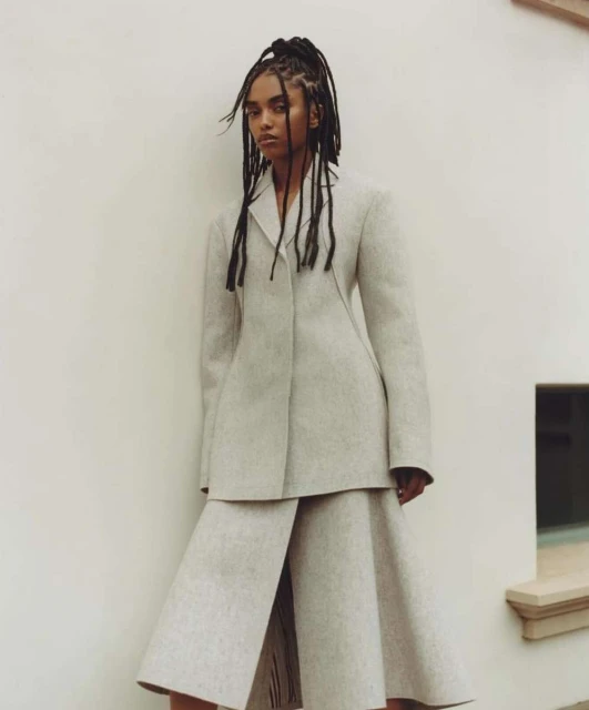 Fashion portrait of a Black woman with braided hair, wearing a neutral blazer and culottes, posing against a textured off-white wall.