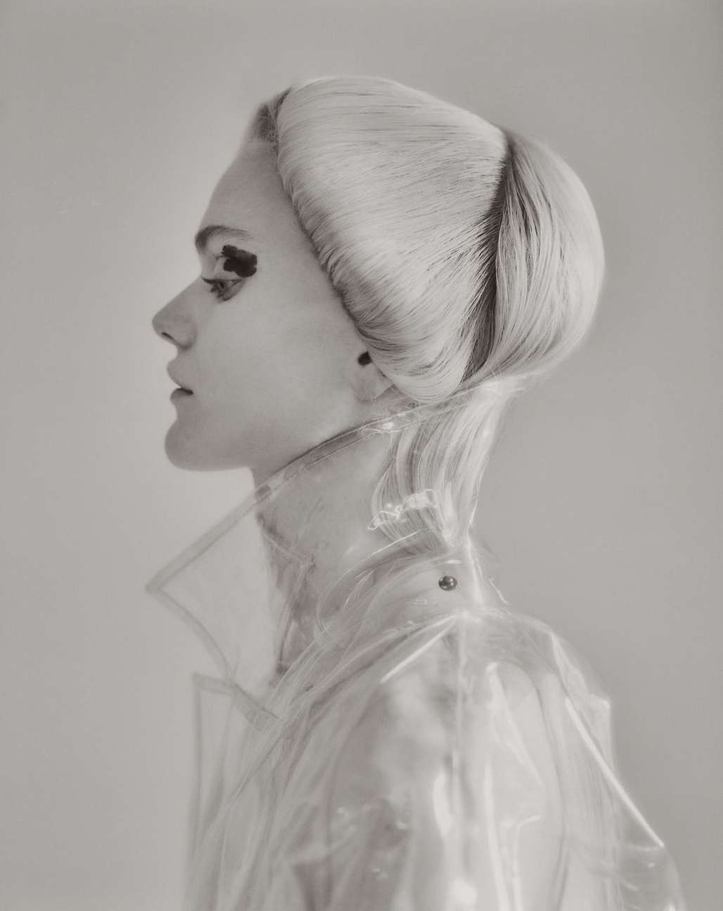 Profile shot of a blonde woman with an elaborate hairstyle wearing a transparent raincoat, set against a neutral gray background.