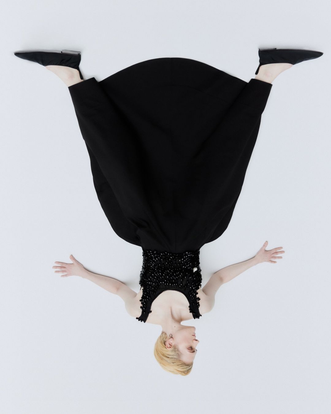 Overhead shot of a woman with blonde hair wearing a black dress and black shoes lying on a white background with her arms outstretched.
