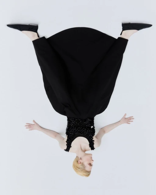 Overhead shot of a woman with blonde hair wearing a black dress and black shoes lying on a white background with her arms outstretched.