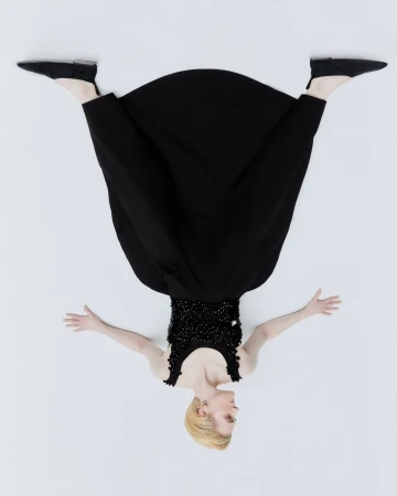 Overhead shot of a woman with blonde hair wearing a black dress and black shoes lying on a white background with her arms outstretched.