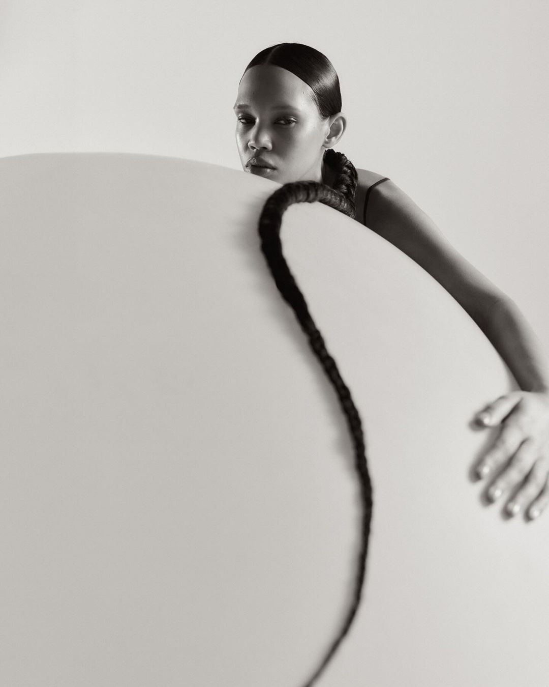 Monochrome portrait of a young woman with a long braid posing behind a circular object in a clean, light-filled studio setting.