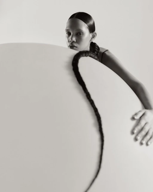 Monochrome portrait of a young woman with a long braid posing behind a circular object in a clean, light-filled studio setting.