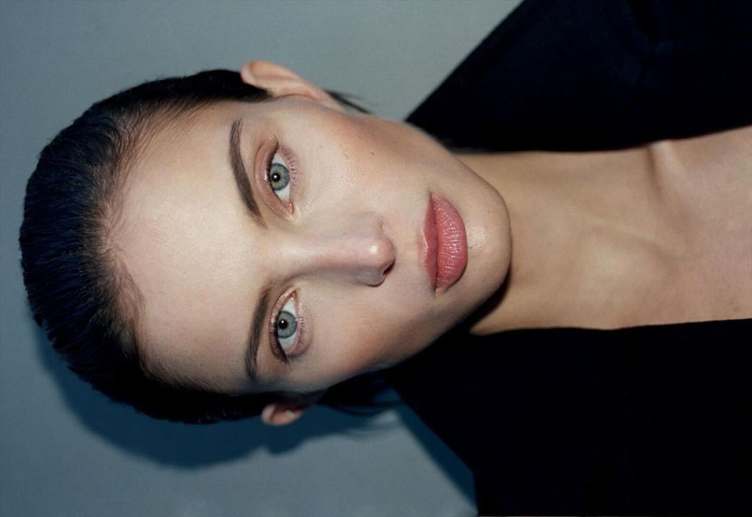 Close-up portrait of a fair-skinned woman with blue eyes and pink lips, wearing a dark top against a grey background. Beauty shot.