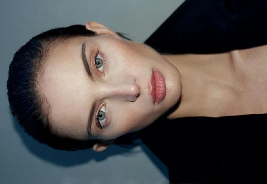 Close-up portrait of a fair-skinned woman with blue eyes and pink lips, wearing a dark top against a grey background. Beauty shot.