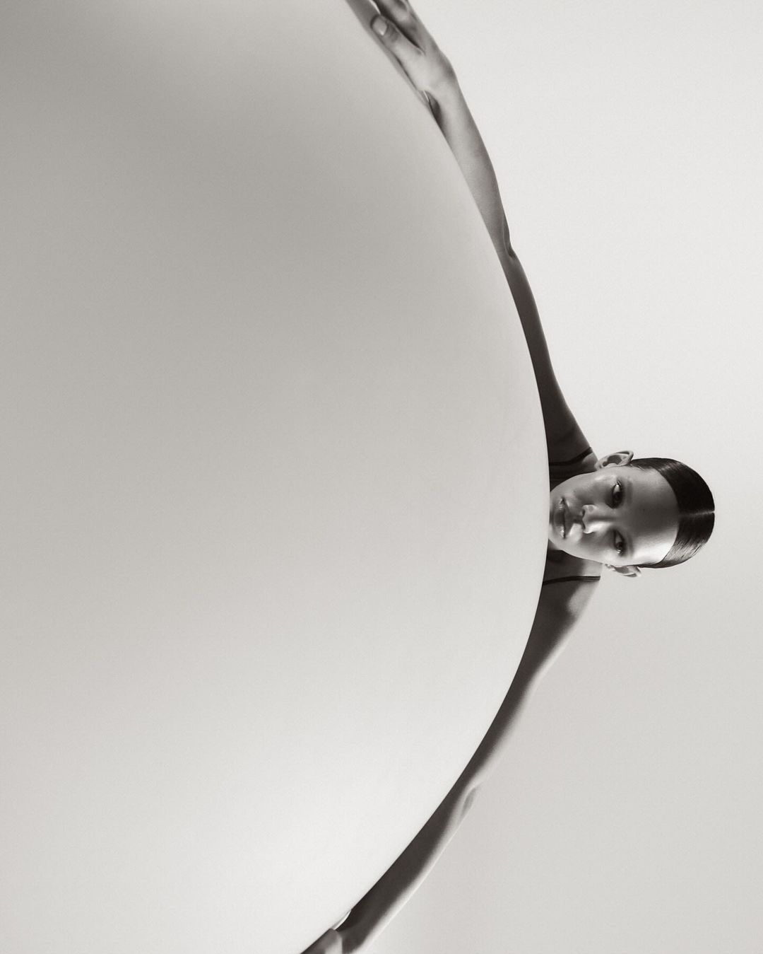 Black and white portrait of a young woman with fair skin and slicked-back dark hair, posing on an off-white abstract background.