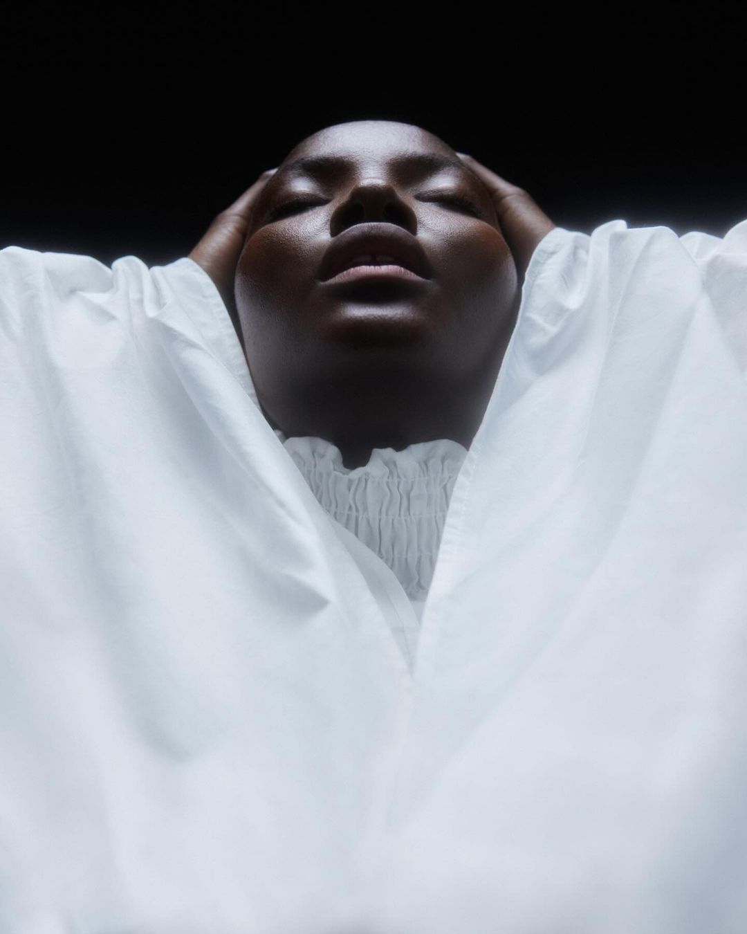 Close-up portrait of a Black woman with eyes closed, hands on her head, wearing a white top against a dark background in a studio setting.