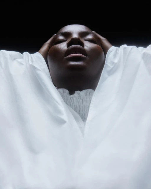 Close-up portrait of a Black woman with eyes closed, hands on her head, wearing a white top against a dark background in a studio setting.