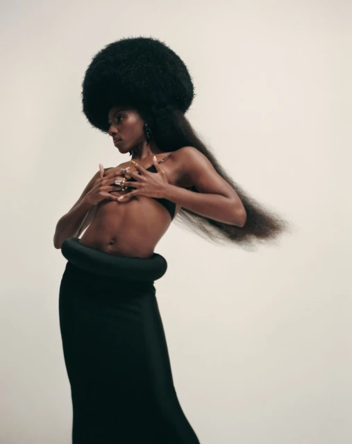 Glamour shot of a Black woman with an afro hairstyle, wearing black skirt and bra in a photo studio, set against a cream backdrop.