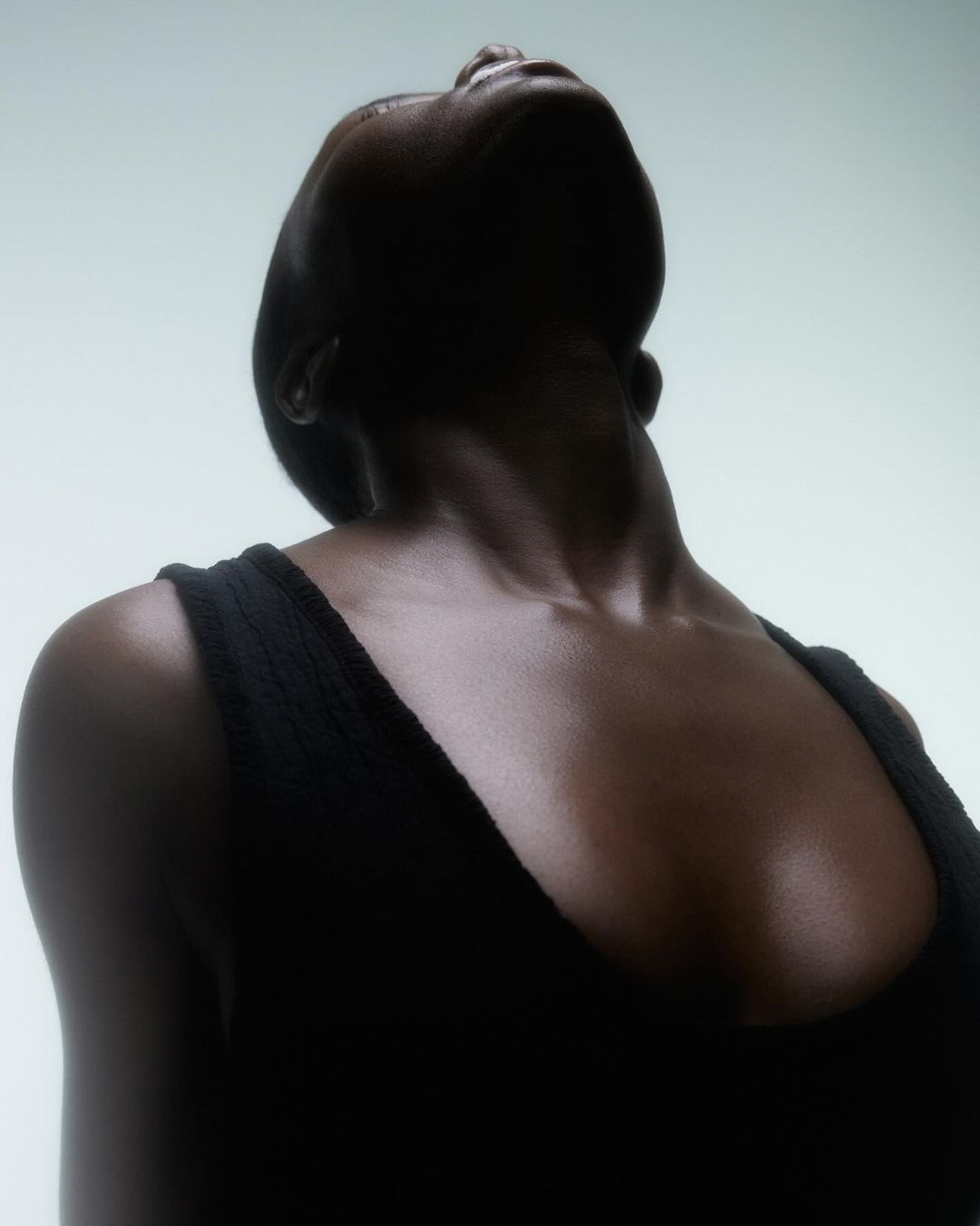 Portrait of an African American woman with dark skin wearing a black tank top, looking up with her head tilted back in a light-filled photo studio.