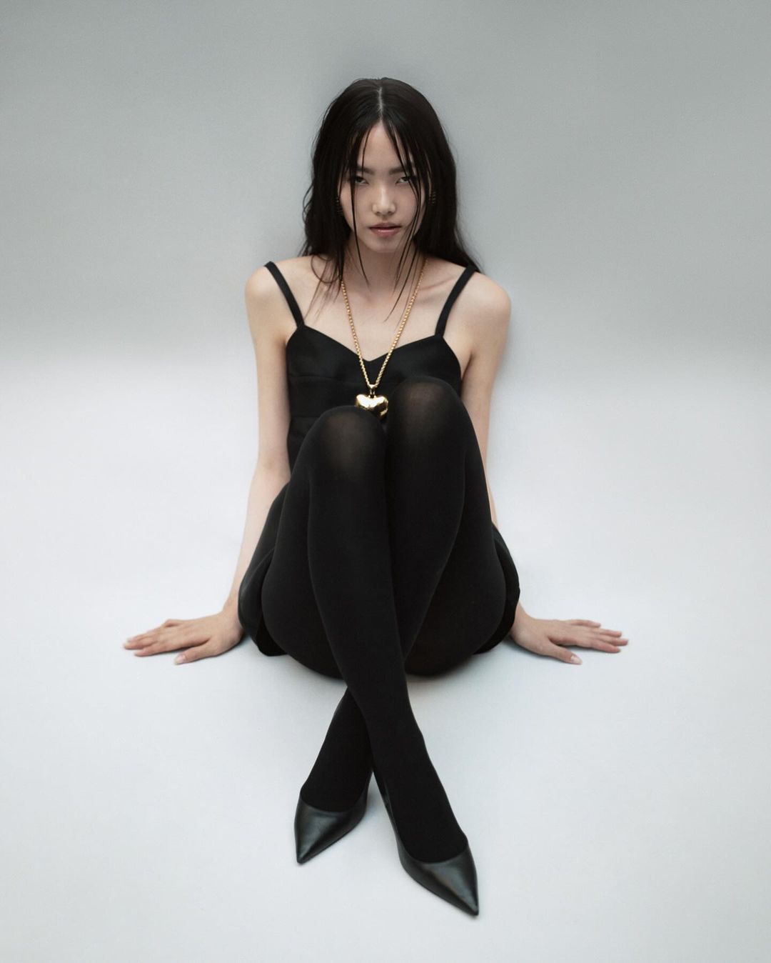 Model with dark hair wearing a black dress and black tights sitting on a grey seamless background with a gold heart necklace