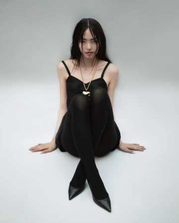 Model with dark hair wearing a black dress and black tights sitting on a grey seamless background with a gold heart necklace