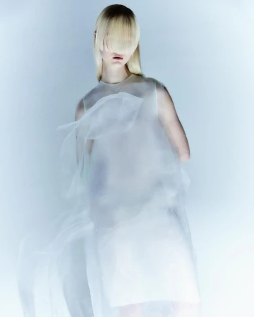Model with long blonde hair covering eyes wearing a sheer white dress against a pale gradient backdrop.