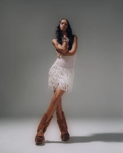 Full-shot of a woman in a fringed dress and platform boots, arms crossed, in a neutral studio. Fashion, style, and portrait photography.