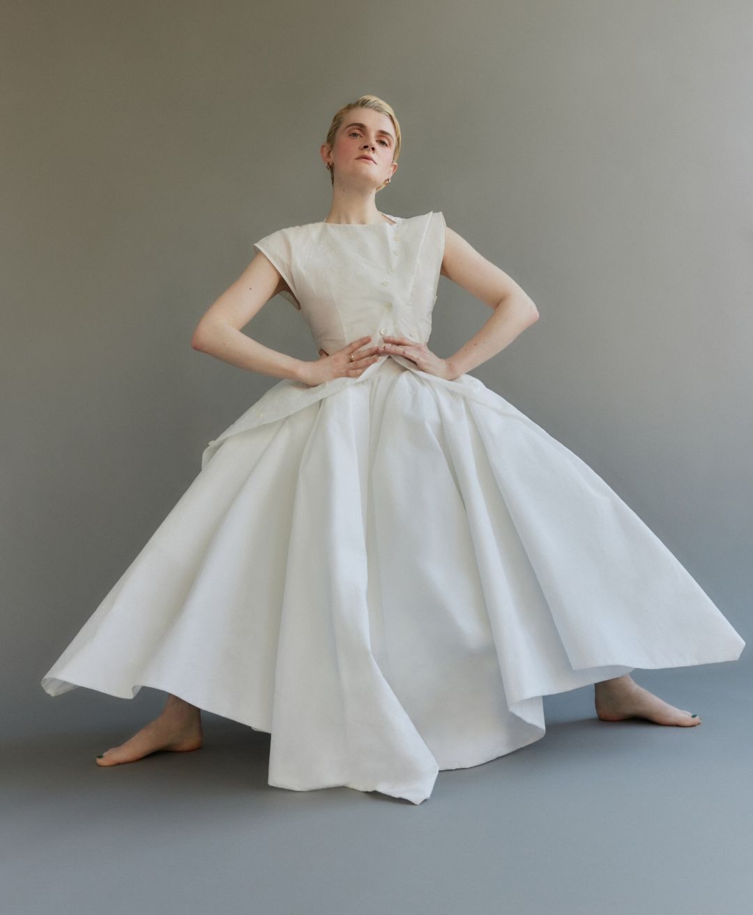 Elegant woman in a sleeveless white top and flared skirt stands in a neutral grey studio, posing with her hands on her hips.
