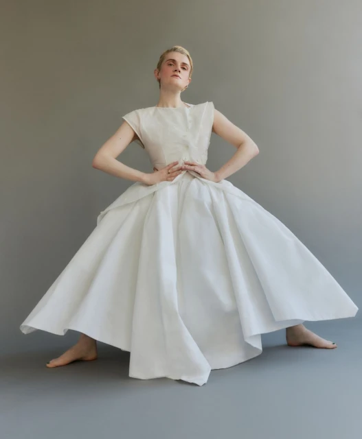 Elegant woman in a sleeveless white top and flared skirt stands in a neutral grey studio, posing with her hands on her hips.