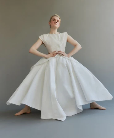 Elegant woman in a sleeveless white top and flared skirt stands in a neutral grey studio, posing with her hands on her hips.