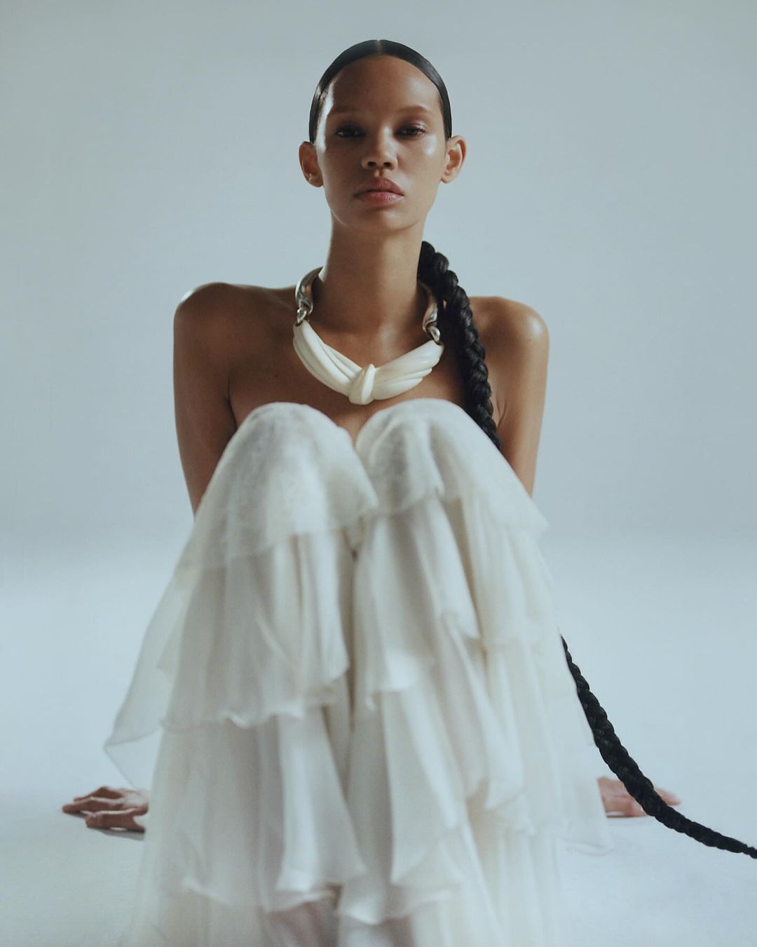 Model with long braided hair wearing a white ruffled dress and chunky necklace against a pale seamless background