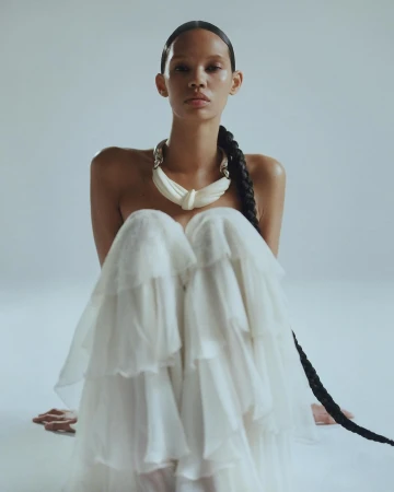 Model with long braided hair wearing a white ruffled dress and chunky necklace against a pale seamless background