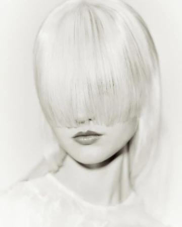 Close-up portrait of a woman with long pale blonde hair covering her eyes and a white top against a light background.