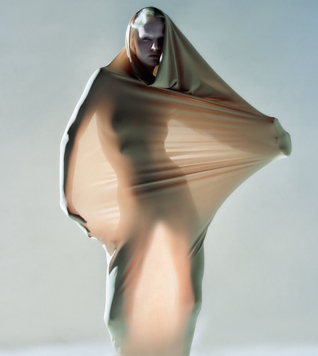 Artistic portrait of a person draped in beige fabric, posing in a studio with soft lighting, showcasing unique draped clothing and style.
